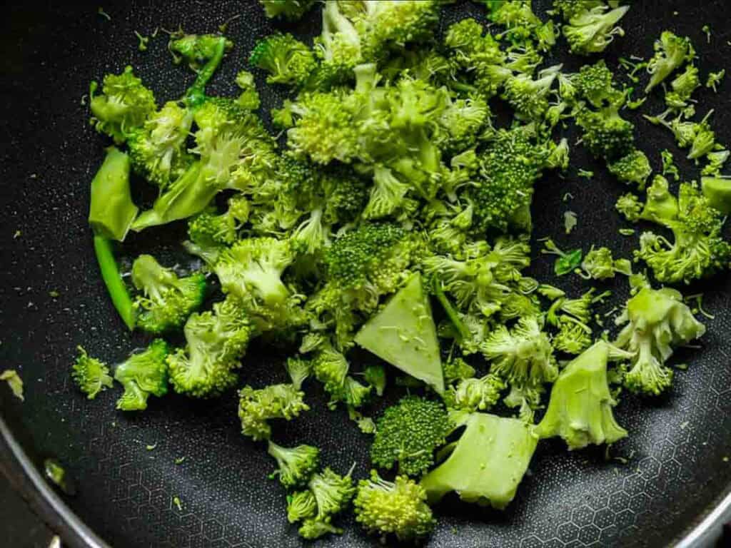 Chopped broccoli florets in a black non-stick frying pan, ready for cooking. The bright green of the broccoli contrasts with the black textured surface of the pan.